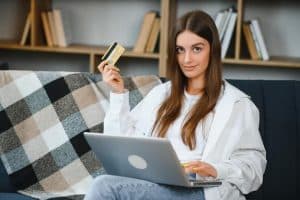 Mulher de cabelos longos sentada em sofá, usando notebook e segurando um cartão de crédito dourado, com expressão confiante em ambiente doméstico.