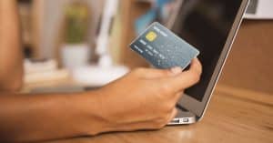 Pessoa usando cartão de crédito em frente a um notebook, simbolizando transações online e uso de tecnologia financeira.